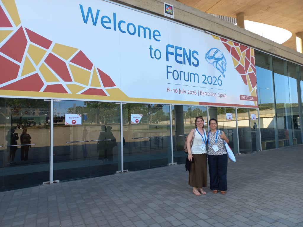 Two conference participants stand in front of the entrance to the FENS Forum 2026 venue beneath a large welcome banner in Barcelona.