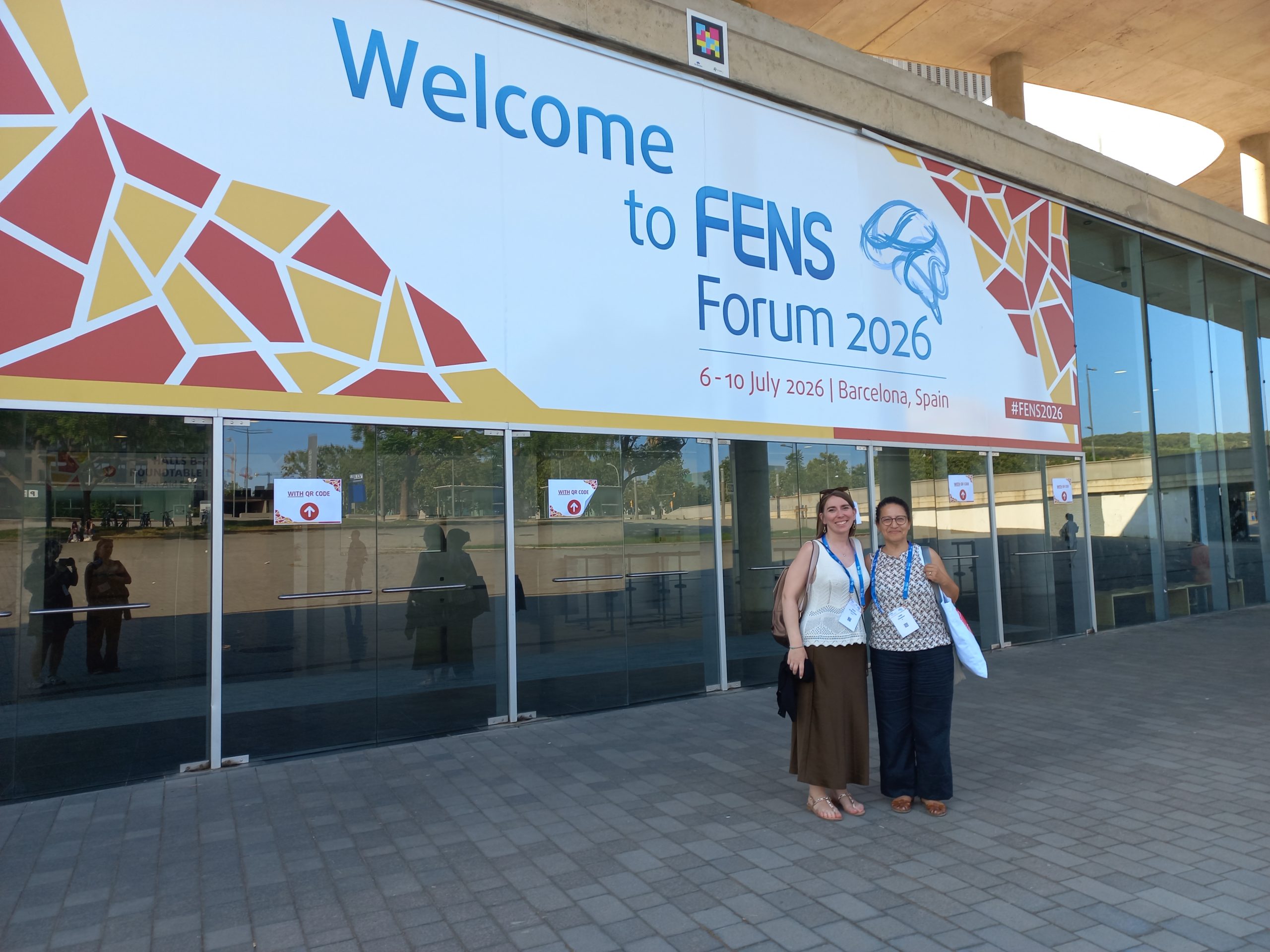 Two conference participants stand in front of the entrance to the FENS Forum 2026 venue beneath a large welcome banner in Barcelona.