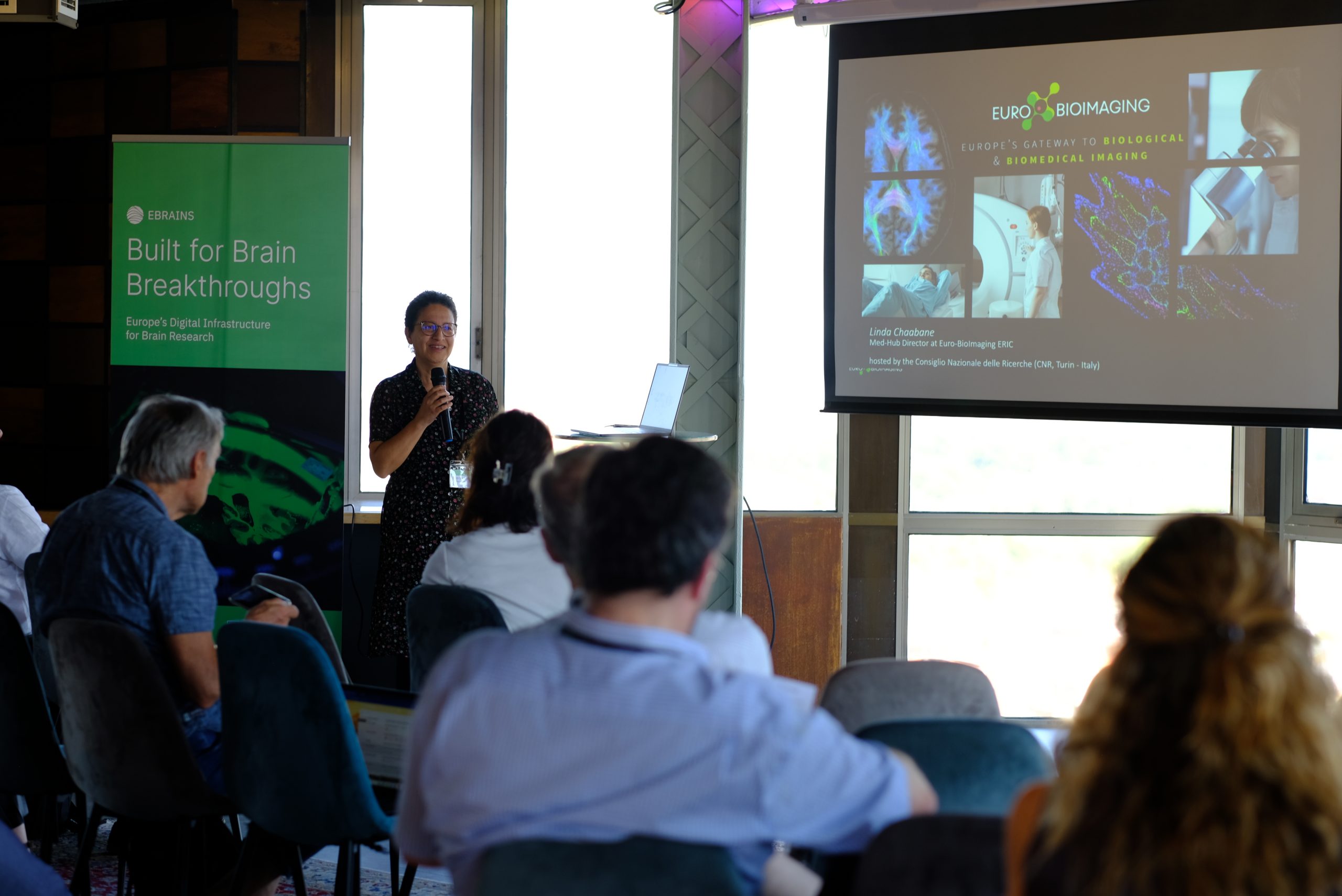 A speaker presents a Euro-BioImaging session to an audience, with an EBRAINS banner displayed beside the presentation screen.